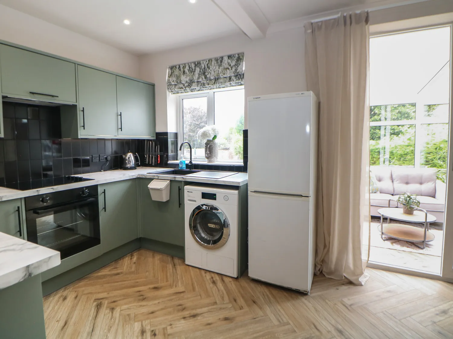 Sage-green shaker kitchen with black tile splashback and herringbone floor